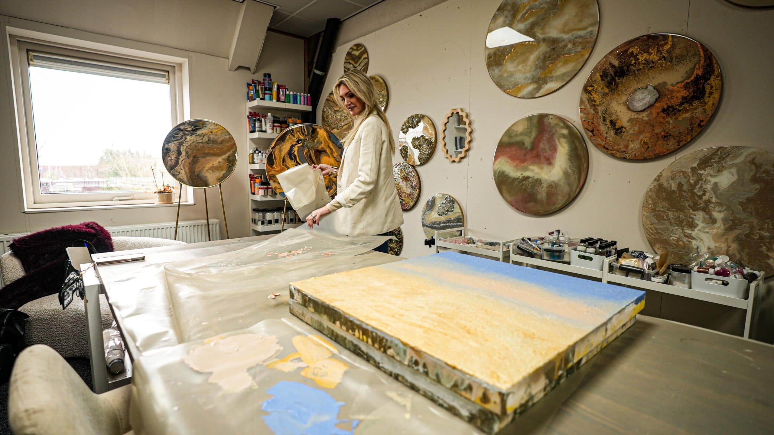Angela in haar atelier aan de werktafel, met een wand vol ronde epoxywerken achter haar